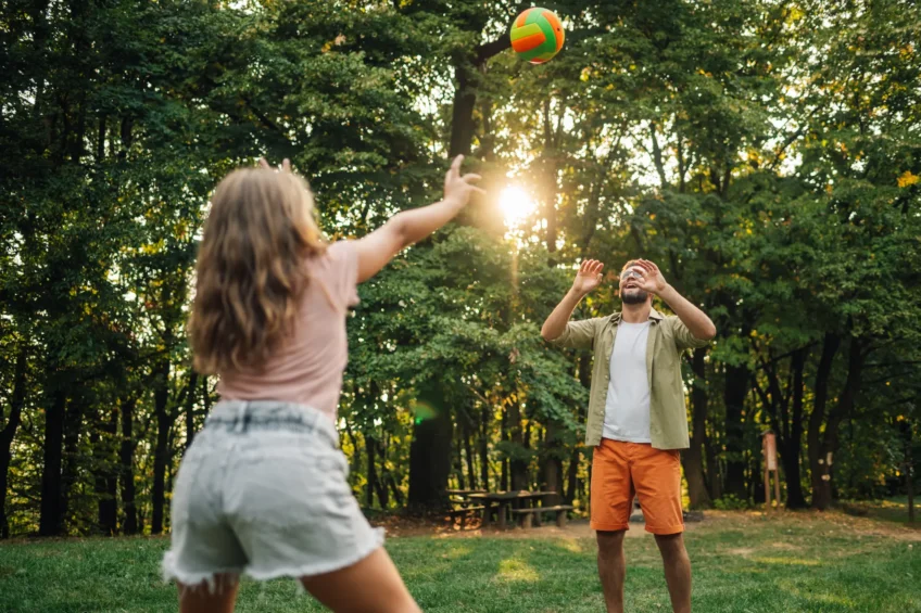 A man and a woman playing volleyball in a sunny forest clearing, smiling and enjoying a casual outdoor game surrounded by tall green trees.