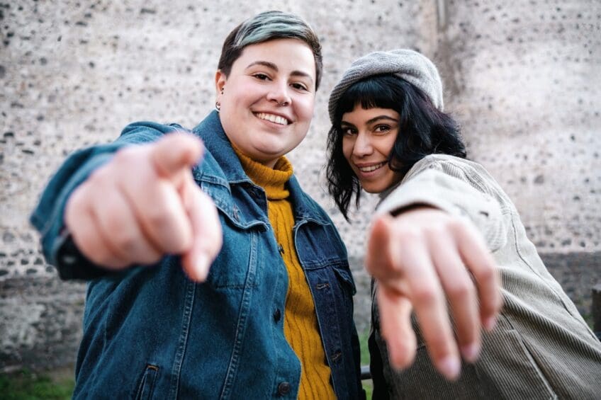 Happy cheerful women pointing at you looking at the camera outdoors against an ancient wall