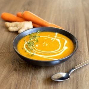 Carrot ginger soup on a wooden countertop next to fresh carrots and ginger