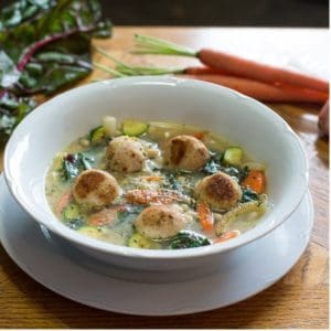 Bowl of turkey meatball and vegetable soup on a white plate next to fresh ingredients