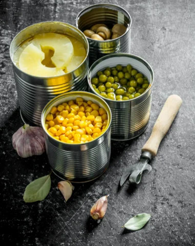 Open cans containing sliced pineapple, mushrooms, green peas, and corn arranged on a dark surface with garlic cloves, bay leaves, and a vintage-style can opener nearby.