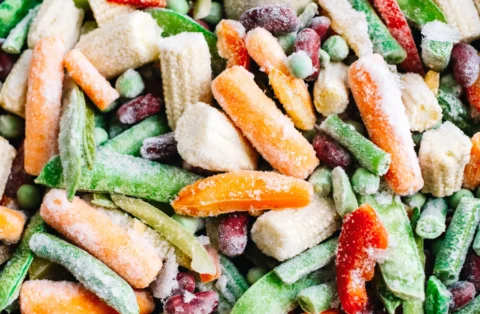 Close-up of a colorful mix of frozen vegetables including carrots, green beans, peas, kidney beans, red peppers, and baby corn, all covered with a layer of frost.