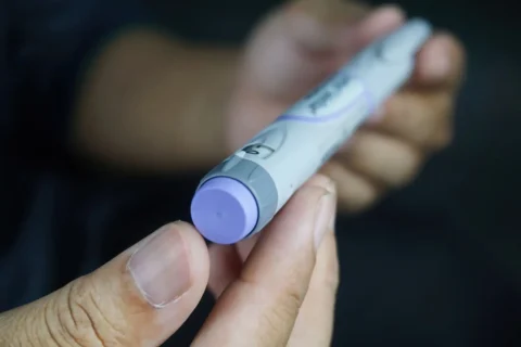 Close-up of a person’s hands holding an insulin pen with a purple cap, commonly used for diabetes management. The background is blurred, focusing attention on the medical device.