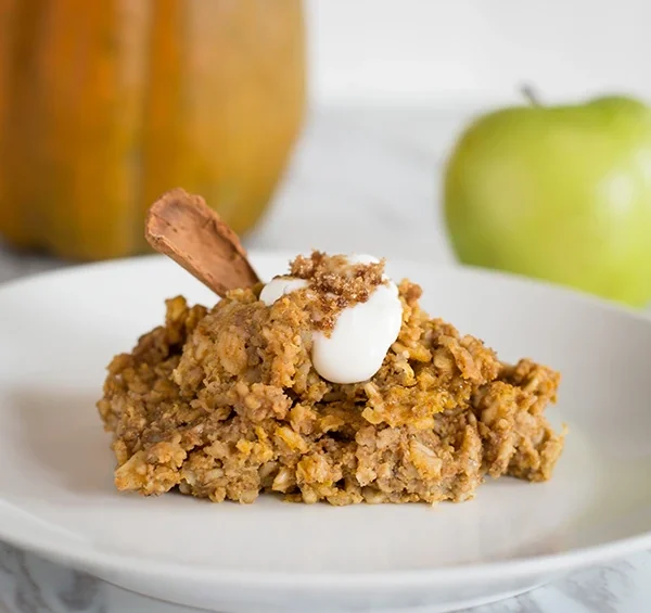 A serving of baked pumpkin oatmeal topped with a dollop of yogurt, a sprinkle of crumble, and a cinnamon stick, plated with a pumpkin and green apple in the background.