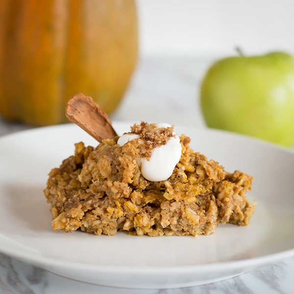 A serving of baked pumpkin oatmeal topped with a dollop of yogurt, a sprinkle of crumble, and a cinnamon stick, plated with a pumpkin and green apple in the background.