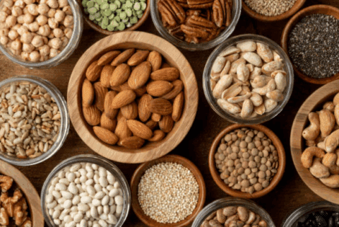 Bowls of assorted nuts, seeds, legumes, and whole grains displayed on a wooden surface.