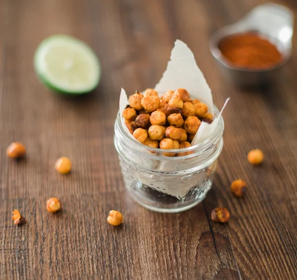 Lime sriracha roasted chickpeas in a small glass jar on a wooden table with lime and spices.