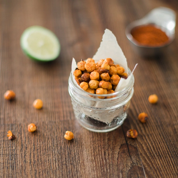 Lime sriracha roasted chickpeas in a small glass jar on a wooden table with lime and spices.