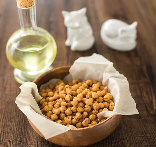 Wooden bowl lined with parchment paper and filled with roasted crispy chickpeas on a rustic wooden table, with a glass bottle of oil and two white ceramic figurines in the background.