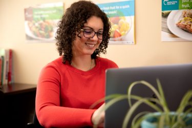 A cheerful woman with curly hair and glasses wearing a red sweater is working on a laptop in an office setting. There's a healthy eating poster visible in the background, promoting fresh fruit as a snack.