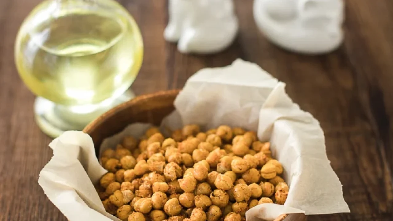 Wooden bowl lined with parchment paper and filled with roasted crispy chickpeas on a rustic wooden table, with a glass bottle of oil and two white ceramic figurines in the background.