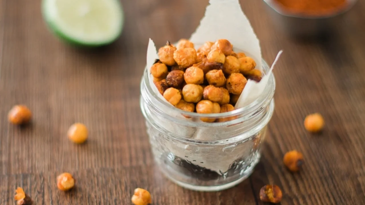 Lime sriracha roasted chickpeas in a small glass jar on a wooden table with lime and spices.