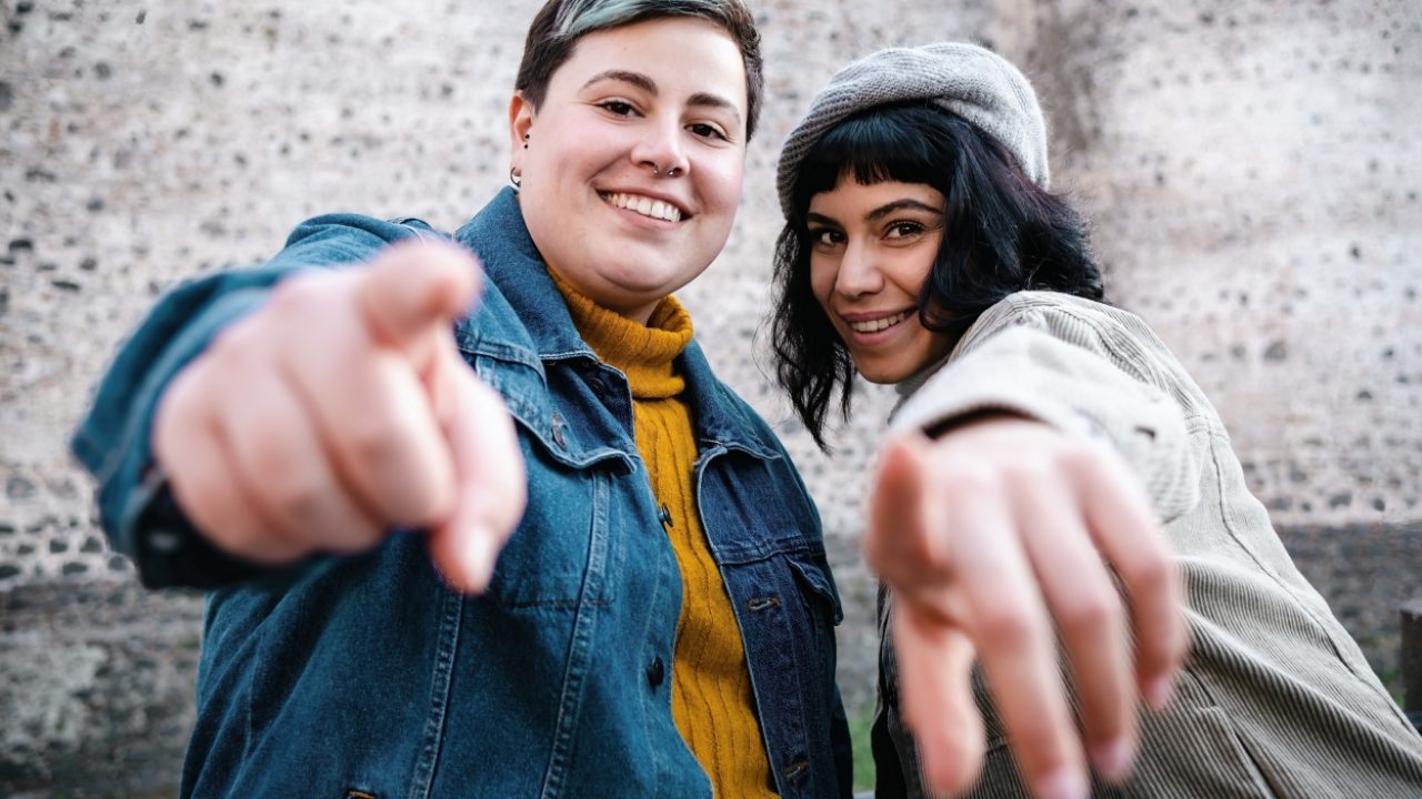 Happy cheerful women pointing at you looking at the camera outdoors against an ancient wall